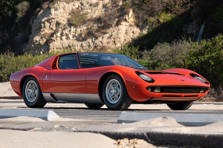 Red Lamborghini Miura P400 parked on a road next to sand in the foreground and a cliffside in the background.