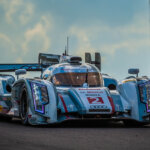 Blue Audi E-tron Quattro hypercar driving on a race course with blue sky and clouds in the background with spectators stands in the background.