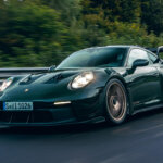 Green Porsche 911 GT3 RS with bronze wheels driving on a racetrack with trees in the background.