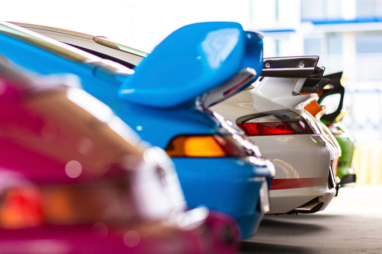 Several Porsche cars all different colors parked next to one another, a purple one out of focus, a blue one, a white one in a showroom floor.