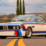 BMW 3.0 CSL "Batmobile" parked on the road with yellow double lane divider going from foreground and underneath the car with houses, trees, and blue sky with clouds in the background.