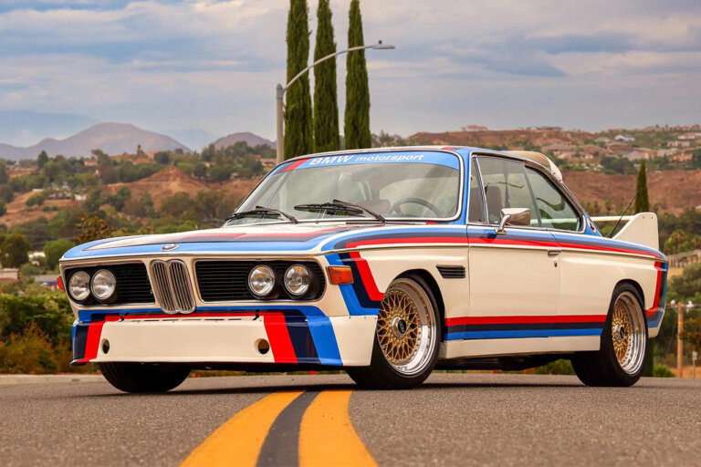 BMW 3.0 CSL "Batmobile" parked on the road with yellow double lane divider going from foreground and underneath the car with houses, trees, and blue sky with clouds in the background.